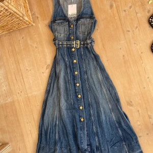 Lång jeansklänning från Ganni - Supersnygg lång jeansklänning från Ganni med ärmlös design, markerad midja och bälte med gulddetaljer. Klänningen har knappar hela vägen framtill, fickor på bröstet och klassisk blå denim-look. Aldrig använd, prislappen sitter kvar. Nypris 4595 på hemsidan. 