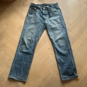 Levi's 501 blå jeans straight fit - Klassiska Levi's 501 jeans i blå tvätt med raka ben och normal passform. Jeansen har fem fickor, knappgylf och den ikoniska läderpatchen bak i midjan. Materialet är slitstarkt denim i bomull och färgen har en snygg, något blekt look. Jeansen har några små revor i benöppningen därav det låga priset.