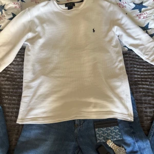 Vit långärmad tröja från Polo Ralph Lauren - Vit långärmad tröja från Polo Ralph Lauren i storlek S. Tröjan är tillverkad i 100% bomull med en snygg, diskret struktur och har den klassiska broderade Polo-loggan i marinblått på bröstet. Perfekt för en clean och stilren look.