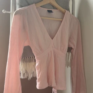 Ljusrosa omlottopp från Gina Tricot - Supersöt ljusrosa omlottopp från Gina Tricot med v-ringning och långa ärmar. Toppen har knytband i midjan och är tillverkad i ett lätt, krinklat tyg som ger en avslappnad känsla. Perfekt för dig som gillar feminina detaljer och en mjuk färgpalett.