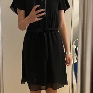 Svart midiklänning från H&M XS - Svart klänning från H&M i storlek XS. Modellen har kort ärm, knytband i midjan och är tillverkad i mjuk polyester. Klänningen är luftig och har en lätt transparent känsla nertill. Perfekt för dig som gillar en enkel men stilren look. Använd en gång.