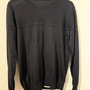 Svart tröja från J.Lindeberg - Snygg svart stickad tröja från J.Lindeberg med diskret logga i nacken. Tröjan har rund halsringning och långa ärmar, perfekt för lager-på-lager. Enkel och stilren design som funkar till det mesta.