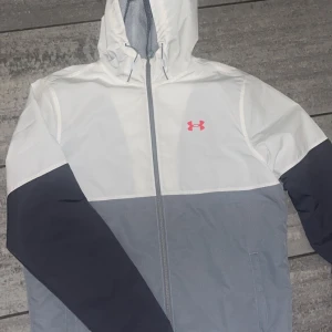 Under Armour vindjacka med huva - Snygg vindjacka från Under Armour i vitt, grått och mörkblått. Jackan har huva med dragsko, hel dragkedja och två sidofickor. Perfekt för blåsiga dagar och sportiga outfits. Under Armour-logga i rosa på bröstet ger en sportig vibe.