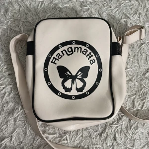 Vit axelväska från Hangmatta - Snygg väska med fjärils tryck på framsidan. Justerbar axelrem och är i fint skick. Perfekt storlek på väska för att ha allt man behöver med sig. Coola detaljer, funkar för killar & tjejer och en fett unik logga.