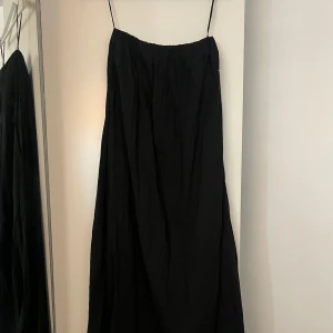Svart långklänning med tunna axelband - En svart långklänning med tunna axelband och avslappnad passform. Klänningen har en enkel och stilren design, perfekt för dig som gillar minimalistiskt mode. Materialet känns lätt och flowy, vilket gör den skön att bära under varma dagar.
