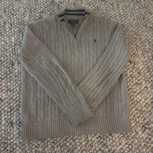 Grå kabelstickad tröja Polo Ralph Lauren - Kabelstickad grå tröja från Polo Ralph Lauren med halv dragkedja och ribbade muddar. Hög krage med marinblå och grå ränder, broderad logga på bröstet och långärmad modell. Perfekt för lager-på-lager under kyliga dagar. Köpt ny av mig. Passar pojke 14-16 år. Nypris ca 1400kr