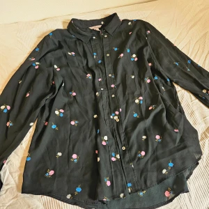 Svart blommig skjorta - Svart skjorta från H&M med färgglada blommor i rosa, blått och vitt. Tunnt och flowy material, lite halvgenomskinlight. Storleken står som 50: jag (36) har haft den som oversized, så vet inte riktigt om den storleken stämmer. Skön på sommaren om man vill skydda sig mot solen ett tag utan att bli för varm. 