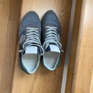 Philippe Model TRPX sneakers grå/grön - Snygga Philippe Model TRPX Low Man sneakers i grå mocka och textil med gröna detaljer vid hälen. Skorna har klassisk snörning, rund tå och vit sula. Perfekta för dig som gillar stilrena och sportiga sneakers med en unik touch.