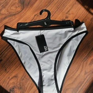 Vit bikiniunderdel från 157 - Snygg vit bikiniunderdel från 157 med svarta kontrastkanter. Klassisk modell med hög midja och rena linjer, perfekt för stranden eller poolen. Tillverkad i stretchigt syntetmaterial som sitter skönt och formar sig efter kroppen.
