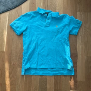 Blå polo t-shirt från Ralph Lauren - Snygg blå polo t-shirt från Ralph Lauren i storlek M. Klassisk piké med korta ärmar, krage och två knappar framtill. Ikonisk broderad logga i orange på bröstet. Tillverkad i mjuk bomull som är skön mot huden. Perfekt för dig som gillar stilren och sportig vibe. Det är även en fläck under loggan som antagligen går att få bort därför prisen.