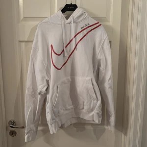 Vit Nike hoodie med röd swoosh - Säljer en vit hoodie från Nike med stor röd swoosh över bröstet och texten 'Just do it.' på axeln. Tröjan har huva med snörning och en stor magficka. Perfekt för dig som gillar sportig och clean stil.