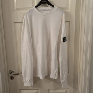 Vit långärmad tröja Calvin Klein - Vit långärmad tröja från Calvin Klein Jeans med svart CK-logga på ärmen. Tröjan har en ribbad struktur och rund halsringning. Perfekt för dig som gillar stilren och enkel design med en touch av street.