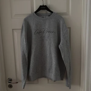 Grå sweatshirt med tryck - Snygg grå sweatshirt med tryck på bröstet där det står 'Café Opera golf'. Tröjan har rund halsringning och långa ärmar, perfekt för en chill och avslappnad stil. Passar bra till jeans eller mjukisbyxor.