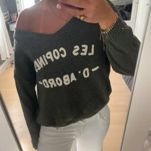 Mörkgrön stickad offshoulder tröja - Säljer en mörkgrön stickad tröja med offshoulder-passning och vit text framtill. Tröjan har en loose fit och långa ärmar, perfekt för dig som gillar en chill och avslappnad stil. Materialet är mjukt och skönt mot huden, och den har en cool, lite croppad look.