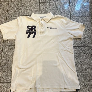 Vit Sail Racing polo SR77 - Vit polo t-shirt från Sail Racing med broderad SR77 i mörkblått och rött på bröstet samt logga på vänster sida. Klassisk krage med tre knappar och korta ärmar. Tillverkad i 100% bomull, perfekt för en chill och sportig look.