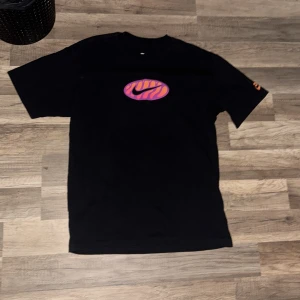 Svart Nike t-shirt med färgglad logga - Svart t-shirt från Nike med korta ärmar och en färgglad logga på bröstet i lila och orange zebra-mönster. Skön passform och klassisk rund halsringning. Perfekt för dig som gillar streetwear och vill sticka ut lite extra.