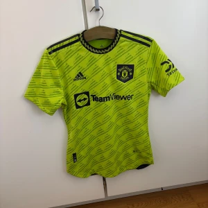 Manchester United Varane matchtröja - Säljer en Manchester United fotbollströja från Adidas i neon-gul färg med svarta detaljer. Tröjan har korta ärmar, klubbmärke på bröstet, sponsorloggor och namn samt nummer 19 för R. Varane på ryggen. Tillverkad i lätt och ventilerande material. Från säsongen 22/23 och går att använda både på sport men även snygg på fritiden!