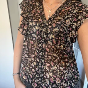 Svart blommig blus med volang - Supersöt svart blus med blommigt mönster i rosa, beige och grönt. Blusen har v-ringning, volangdetaljer vid axlarna och är ärmlös. Perfekt för hösten