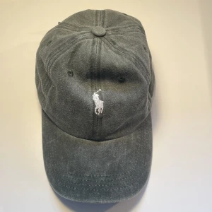 Grå keps från Polo Ralph Lauren - Snygg grå keps från Polo Ralph Lauren med klassisk böjd skärm och justerbart spänne bak. Kepsen har den ikoniska vita broderade polospelaren framtill och är tillverkad i mjuk bomull. Perfekt accessoar för en chill och sportig look.