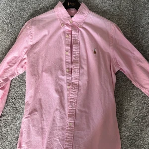 Ralph Lauren - Säljer en rosa skjorta från Polo Ralph Lauren i slim fit med klassisk button down-krage och broderad logga på bröstet. Skjortan är långärmad och tillverkad i bomull, perfekt för en clean och stilren look. Nyskick