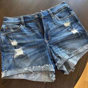 Blå slitna jeansshorts från Hollister - Snygga blå jeansshorts från Hollister med slitna detaljer och fransig kant. Klassisk femficksmodell med dragkedja och knapp.  Shortsen har en normal passform och är tillverkade i mjukt jeanstyg.