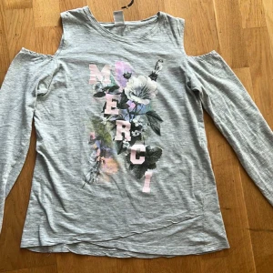 Grå långärmad topp med blommor - Grå långärmad topp med coola cut-out axlar och tryck i pastellfärger. Framsidan har stora rosa bokstäver som bildar ordet 'MERCI' och ett blommigt motiv i vitt, grönt och lila. Mjuk och skön, perfekt för dig som gillar en chill men ändå snygg stil.