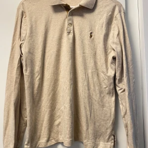 Beige långärmad pikétröja Polo Ralph Lauren - Säljer en beige långärmad pikétröja från Polo Ralph Lauren i slim fit. Tröjan har klassisk krage, två knappar och den ikoniska broderade loggan på bröstet. Mjuk bomullskvalitet som är skön mot huden och passar perfekt till jeans eller chinos.