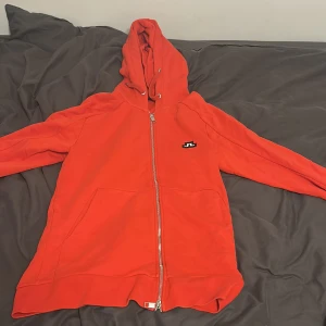 Orange hoodie från J.Lindeberg - Säljer en snygg orange hoodie från J.Lindeberg i storlek L. Tröjan har dragkedja hela vägen, huva med snörning och två stora fickor framtill. Perfekt för chill dagar och har en stilren logga på bröstet. mycket bra skick!