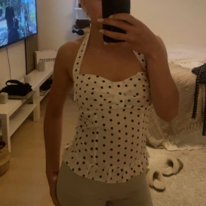 Vit prickig halterneck topp - Supersöt vit halterneck topp med svarta prickar och volangkant nertill. Sitter tajt och har en feminin form med söt knytdetalj vid bysten. Perfekt för dig som gillar retrovibbar och vill sticka ut lite extra.
