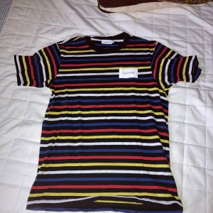 Randig t-shirt från Calvin Klein - Snygg t-shirt från Calvin Klein med färgglada ränder i svart, blått, ratty, gult och vitt. Klassisk passform med rund hals och korta ärmar. Stor logga framtill. Perfekt för dig som vill sticka ut lite extra.