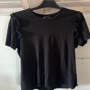 Svart volang-topp från Zara - Svart topp från Zara i storlek L med korta ärmar och snygga volangdetaljer längs axlarna. T-shirten har en klassisk rund halsringning och är gjord i mjuk bomull, perfekt för en stilren och trendig look.