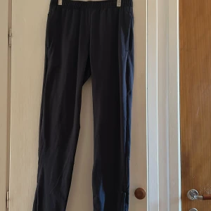 Svarta träningsbyxor med dragkedjor - Svarta träningsbyxor med elastisk midja och dragkedjor både vid fickor och benslut. Snygg och enkel design i syntetmaterial som passar perfekt till träning eller chill. Byxorna har en normal passform och är lätta att röra sig i.