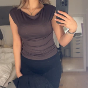 Mörkbrun ärmlös topp med drapering - Säljer en mörkbrun ärmlös topp med snygg draperad halsringning. Toppen har en tight passform och är tillverkad i ett mjukt, stretchigt material som sitter skönt mot kroppen. 