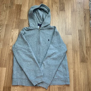 Grå hoodie från Polo Ralph Lauren - Klassisk grå hoodie från Polo Ralph Lauren med dragkedja framtill och broderad marinblå logga på bröstet. Tröjan har huva, långa ärmar och två fickor framtill. Perfekt för en chill och stilren look.