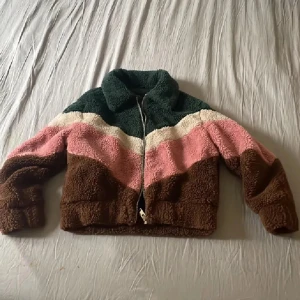Färgglad teddyjacka med dragkedja - Säljer en mysig teddyjacka med coola färgblock i mörkgrönt, beige, rosa och brunt. Jackan har en bred krage och hel dragkedja framtill. Perfekt för dig som gillar retrovibbar och vill sticka ut med en unik look. Materialet är fluffigt och skönt, passar när du vill vara extra cozy.