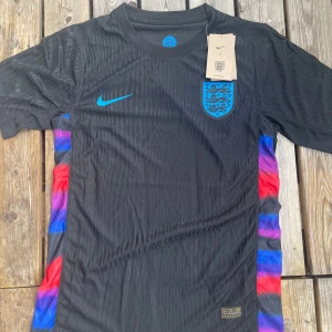 England svart fotbollströja Nike - Snygg svart England fotbollströja från Nike med blått landslagsemblem och logga på bröstet. Sidorna har coola inslag av röd, blå och lila färg. Tröjan är kortärmad och gjord i lätt, ventilerande material som passar perfekt till träning eller match.
