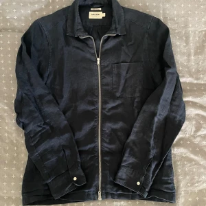 Mörkblå linné overshirt från East West - Snygg mörkblå overshirt från East West i 100% linne. Jackan har en rak passform, bröstficka, klassisk krage och hel dragkedja framtill. Perfekt att slänga över en t-shirt för en clean och avslappnad look.