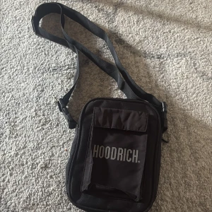 Svart axelväska från Hoodrich - Säljer en svart axelväska från Hoodrich med justerbar rem och praktiskt ytterfack med lock. Väskan har dragkedja upptill och tydlig logga framtill. Perfekt storlek för att bära med det viktigaste. Knappt använd. Köpt från Jd 