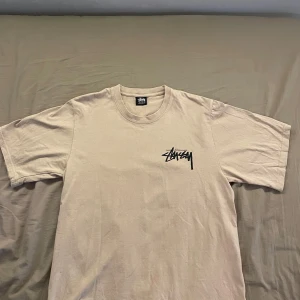 Beige Stüssy t-shirt med lila träningar - Snygg beige t-shirt från Stüssy med svart logga på bröstet och stort tryck på ryggen med lila tärningar. Klassisk passform och rund halsringning. Perfekt för dig som gillar streetwear och vill sticka ut med coola detaljer. Använt 2,3 gånger och den har mycket bra skick. Storlek S, män. Pris kan diskuteras. Skickas inom 1-3 dagar.