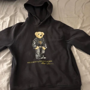 Svart hoodie Polo Bear Ralph Lauren - Svart hoodie från Polo Ralph Lauren med ikoniskt Polo Bear-tryck på bröstet. Klassisk huva, långärmad och normal passform. Mjuk insida och coolt motiv med björn i skinnjacka. Perfekt för dig som gillar streetstyle och vill sticka ut.   Det finns äkthetsbevis !! 
