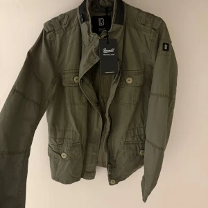 Olivgrön vindjacka från Brandit - Snygg olivgrön vindjacka från Brandit med flera fickor framtill, axelklaffar och coola detaljer. Jackan har dragkedja och knappar, samt en patch med B-logga på ärmen. Perfekt för dig som gillar en avslappnad och urban stil. Perfekt nu till hösten. Jackan är helt ny, pris kan dirkuters vid snabb affär.