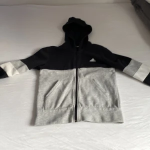 Svart och grå hoodie från Adidas - Snygg hoodie från Adidas i svart och ljusgrått med vit logga på bröstet. Tröjan har dragkedja, huva och stora fickor framtill. Ärmarna har en cool blockdesign i svart, grått och vitt. Perfekt för chill eller träning.