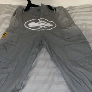 Grå cargopants från cortiez - Säljer ett par grå cargopants från Cortiez med flera stora fickor på sidorna och bak. Byxorna har tryck och broderade loggor samt gula patchar. De är tillverkade i bomull och har en loose fit med justerbar midja. storlek S 