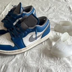 Nike Air Jordan 1 Low blå/vit/grå - Nike Air Jordan 1 Low sneakers i blått, vitt och grått. Klassisk siluett med grå swoosh, blå snörning och perforerad tå. Ovandel i skinn och syntet, lågt skaft och platt sula. Box finns med och creasskydden ingår i priset🙌 bara att skriva om frågor eller om man vill diskutera pris!