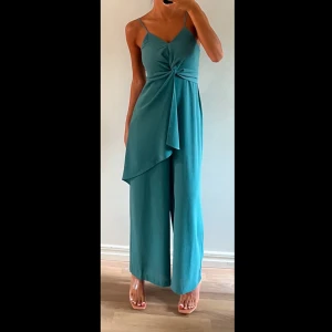 Turkos byxdress med knut och vida ben - Snygg turkos byxdress/jumpsuit med smala axelband, v-ringning och en cool knutdetalj framtill. Vida ben och asymmetrisk omlott-detalj som ger en trendig look. Perfekt för dig som vill sticka ut med stilren vibe.🩵