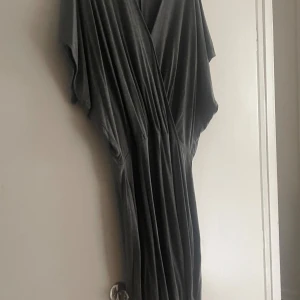 Mörkgrå omlottklänning från Ellos - Snygg långklänning i mörkgrått från Ellos med omlott-design och v-ringning. Klänningen har lös passform, korta ärmar och är tillverkad i mjuk bomullsmix. Perfekt för dig som gillar en avslappnad men stilren look.