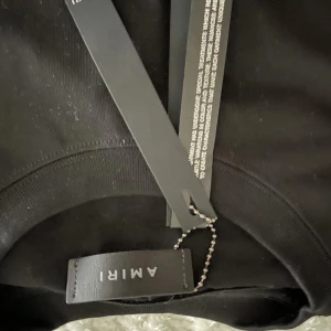 Svart t-shirt från AMIRI med blå logga - Svart t-shirt från AMIRI med stor blå logotyp framtill. Klassisk rund halsringning och korta ärmar. Tillverkad i mjuk bomull som känns skön mot huden. Perfekt för dig som gillar streetwear och vill sticka ut med en snygg logga.