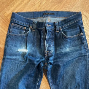 Grim Tim jeans från Nudie Jeans - Säljer ett par blå  jeans från Nudie Jeans med klassisk femficksdesign och snygga slitningar. Jeansen har orangea sömmar, läderpatch bak och en lagad detalj på vänster lår. Cool faded effekt på denimtyget. Storlek 31:34