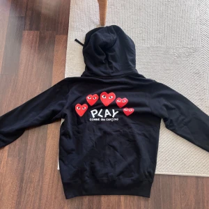 Svart Comme des Garçons PLAY hoodie - Svart hoodie från Comme des Garçons PLAY med dragkedja, huva och fickor. Framsidan har ett rött hjärta med ögon, och på ryggen finns flera röda hjärtan samt PLAY-loggan. Tillverkad i mjuk bomull, perfekt för chill dagar. Långärmad och normal passform.