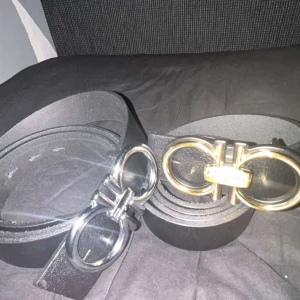 Ferragamo bälten  - Två snygga svarta bälten med bred design och stilrena metallspännen, ett i silver och ett i guld. Perfekt för att lyfta din outfit med en cool detalj. Bältena är gjorda i skinn och har en modern look med dubbla ringar på spännet.